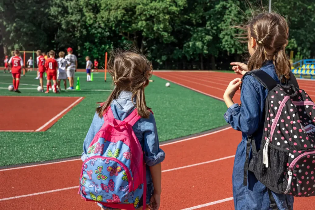 Two children standing near a running track, watching others play